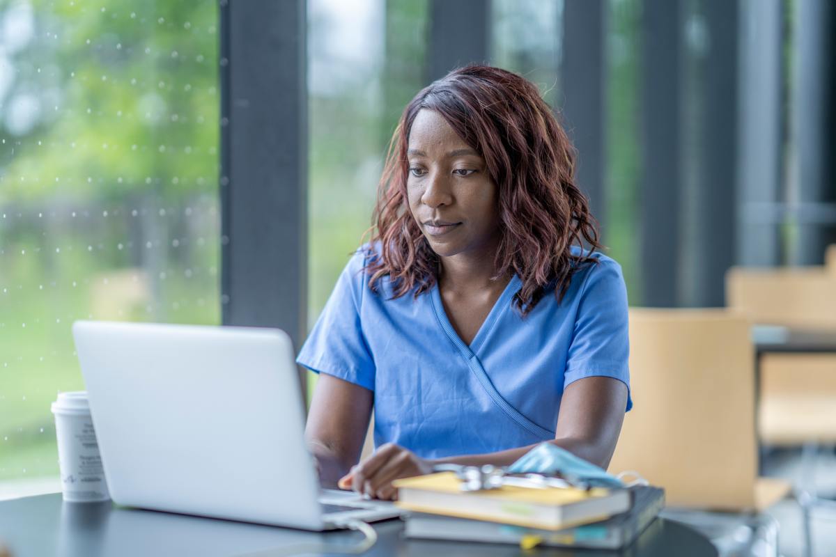 Black female Rochester Regional Health team member studying on a laptop sitting at a cafeteria table with a textbook and notebook